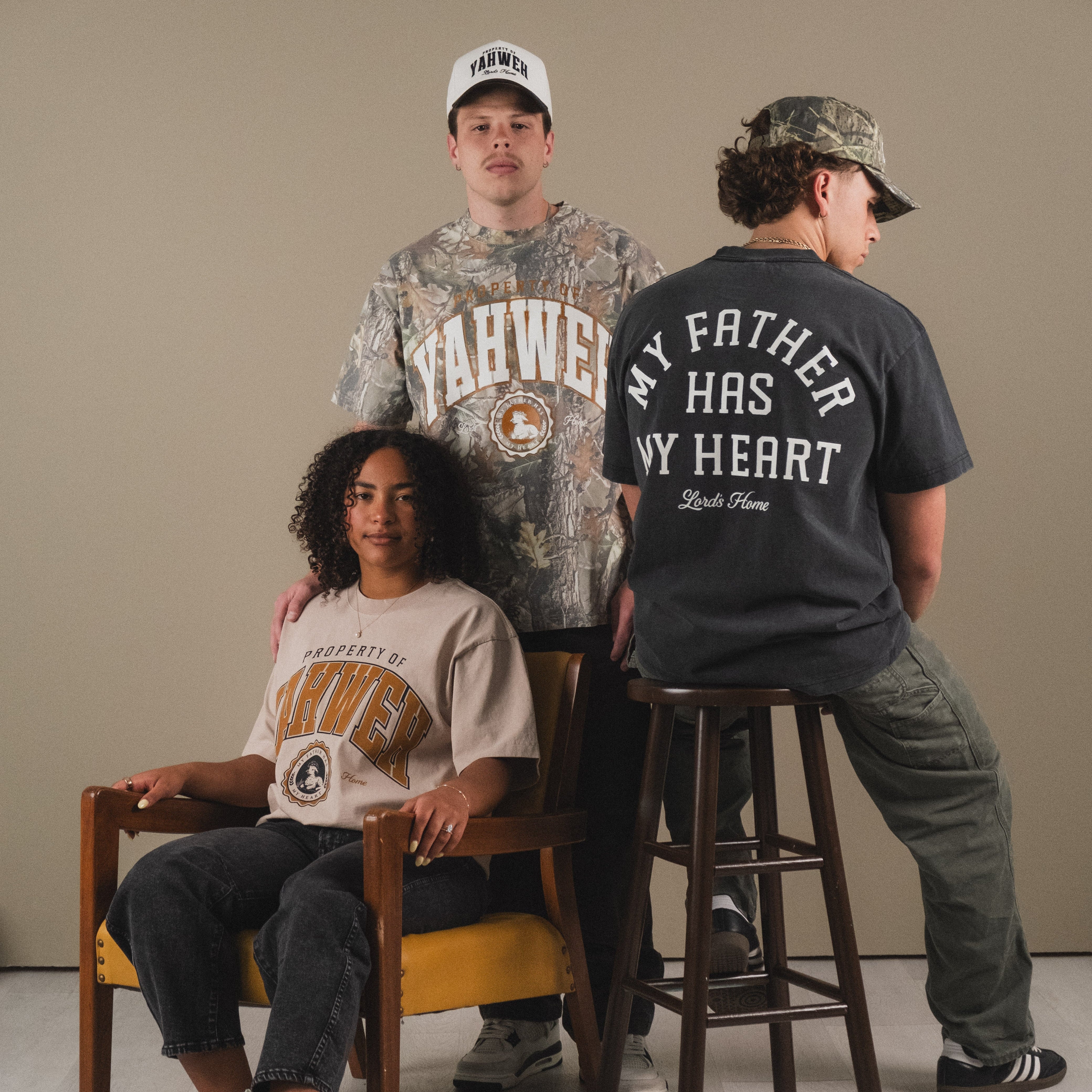 Three models display different graphic t-shirts in a studio setting. One model sits on a wooden chair wearing a beige graphic tee, one stands behind wearing a camouflage patterned tee, and one stands on a stool showing the back of a dark gray graphic tee that reads 'MY FATHER HAS MY HEART'.